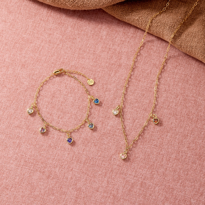 A gold-toned jewelry set featuring a delicate necklace and matching bracelet, both adorned with small clear and blue gemstones, displayed on a textured pink fabric background.